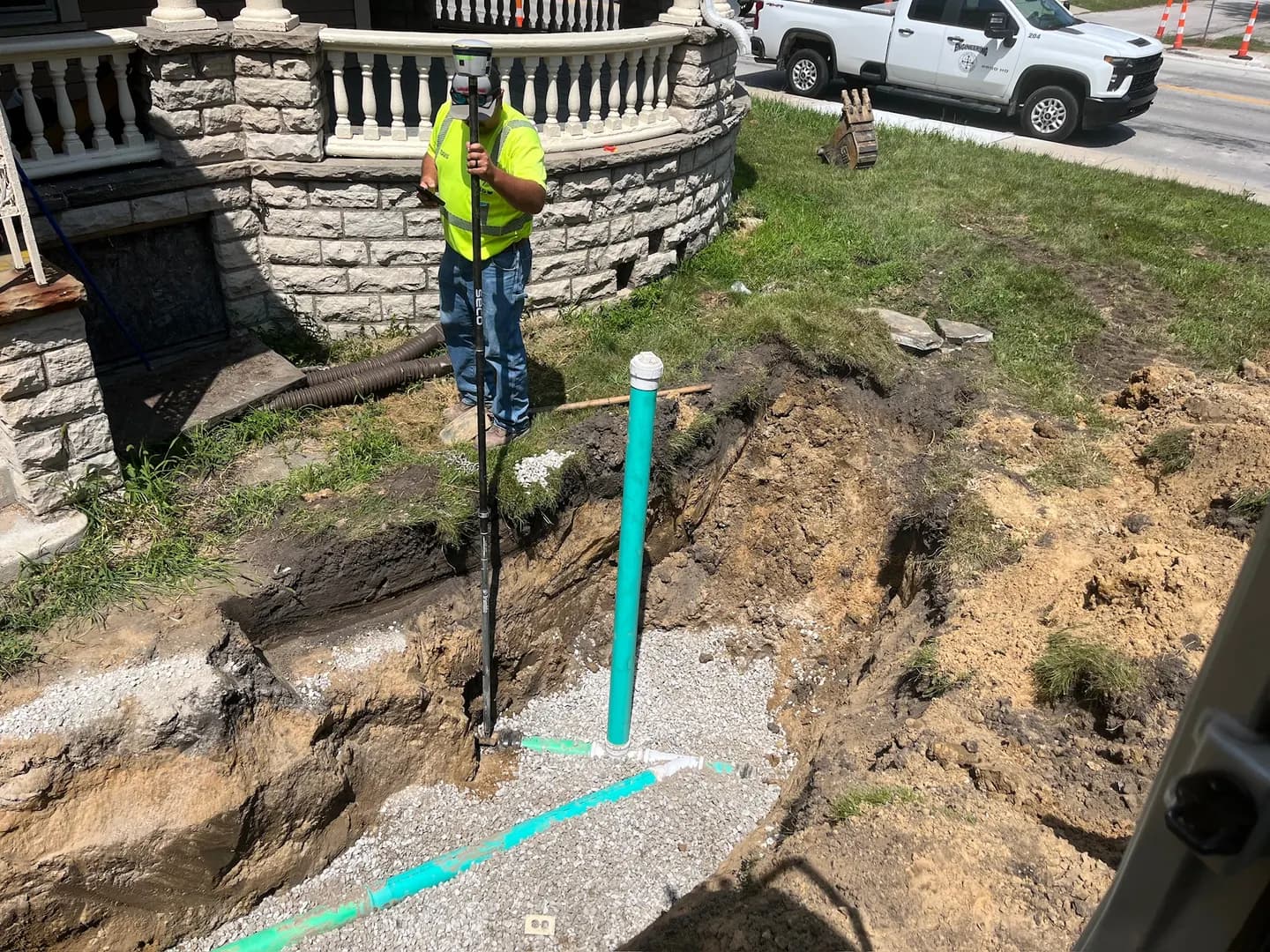 Worker using a survey pole beside green sewer pipe and a cleanout in an open trench